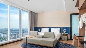 A modern hotel room with a large bed, a small table, a nightstand, and a view of the city. - Novotel Jakarta Pulo Mas (Jakarta)