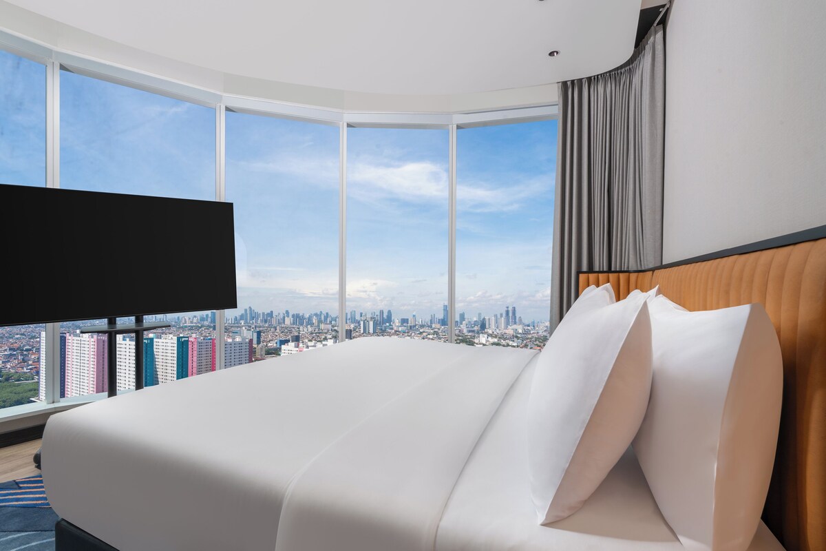 Ein modernes Hotelzimmer mit einem großen Bett, einem Flachbildfernseher und Blick auf die Stadt durch große Fenster.