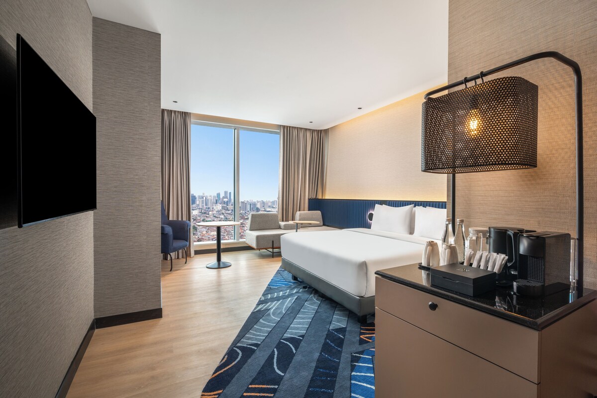 Ein Hotelzimmer mit einem großen Bett, einem Schreibtisch mit Kaffeemaschine, einem Fernseher und Blick auf die Stadt.