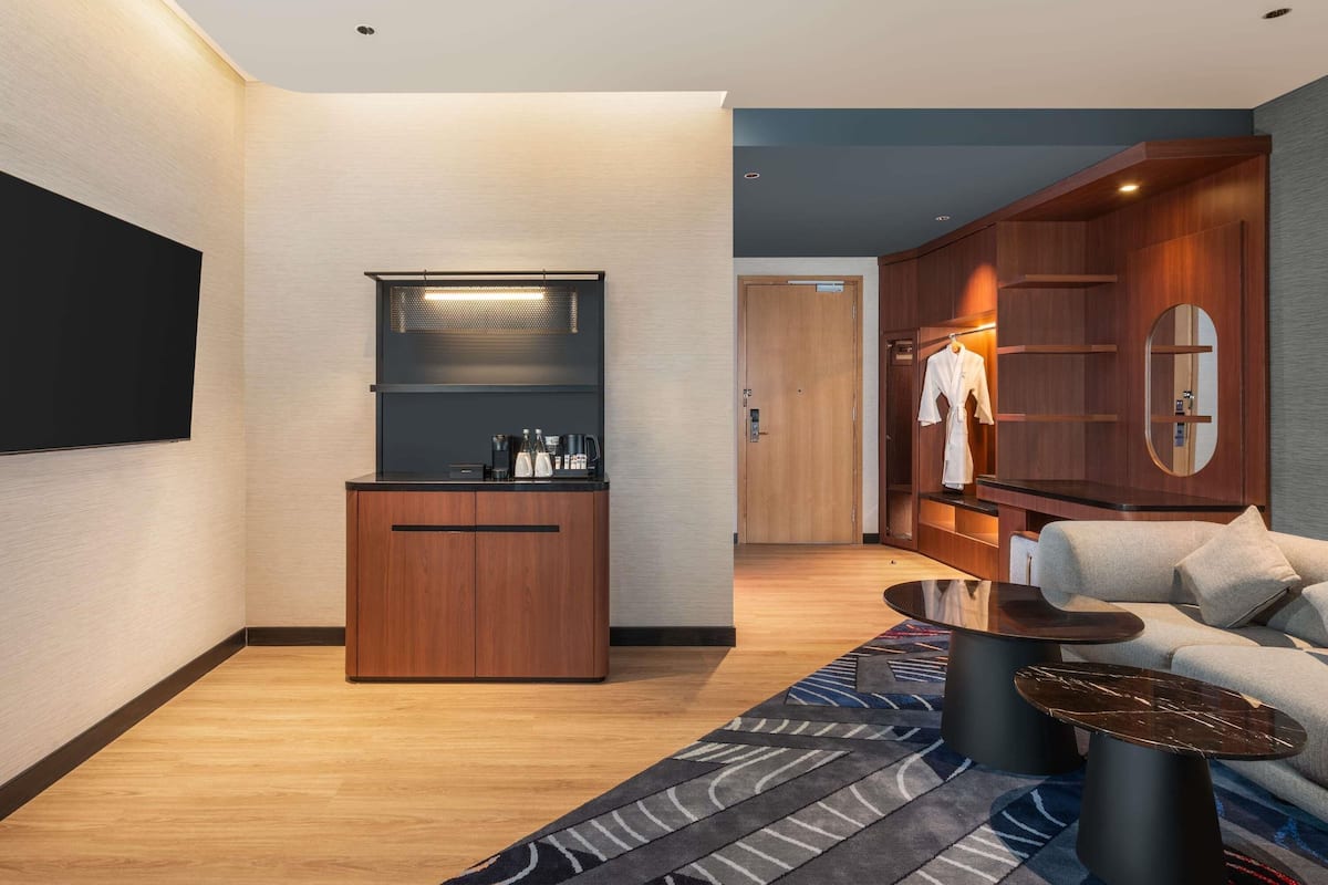 Una habitación de hotel moderna con un mueble de madera, un televisor de pantalla plana, una zona de estar con un sofá y una mesa de centro, y una bata colgada en un armario de madera.