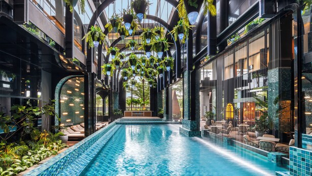 An indoor swimming pool with a tiled edge, surrounded by seating areas and hanging plants. - Novotel Tangerang Bsd City (Pagedangan)