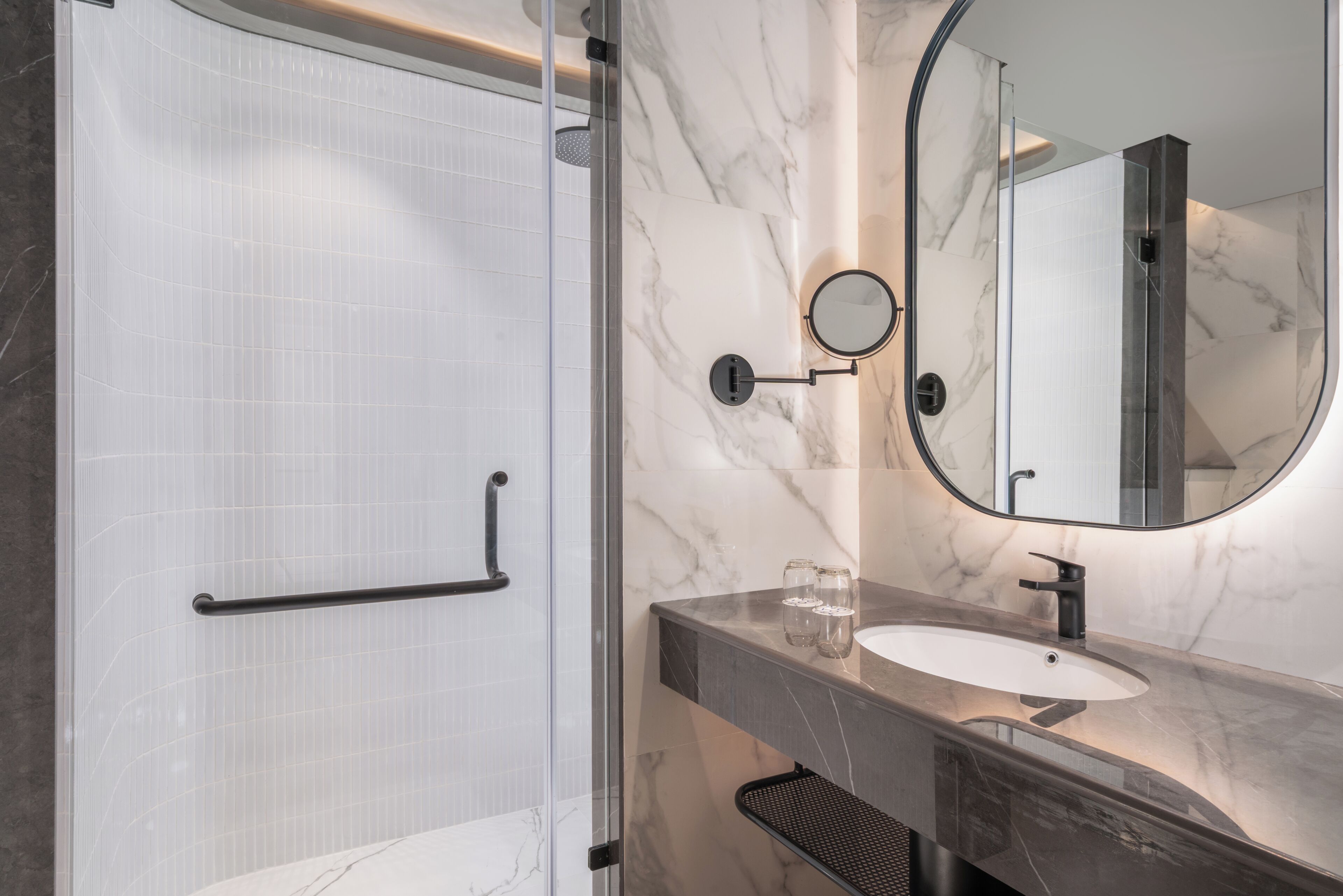 A modern bathroom with a glass shower enclosure, a marble-tiled wall, a round mirror, and a sink with a black faucet.