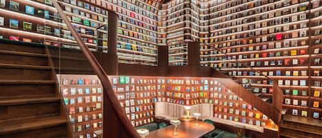 A library with bookshelves on all sides, a central staircase, and a reading area with chairs.