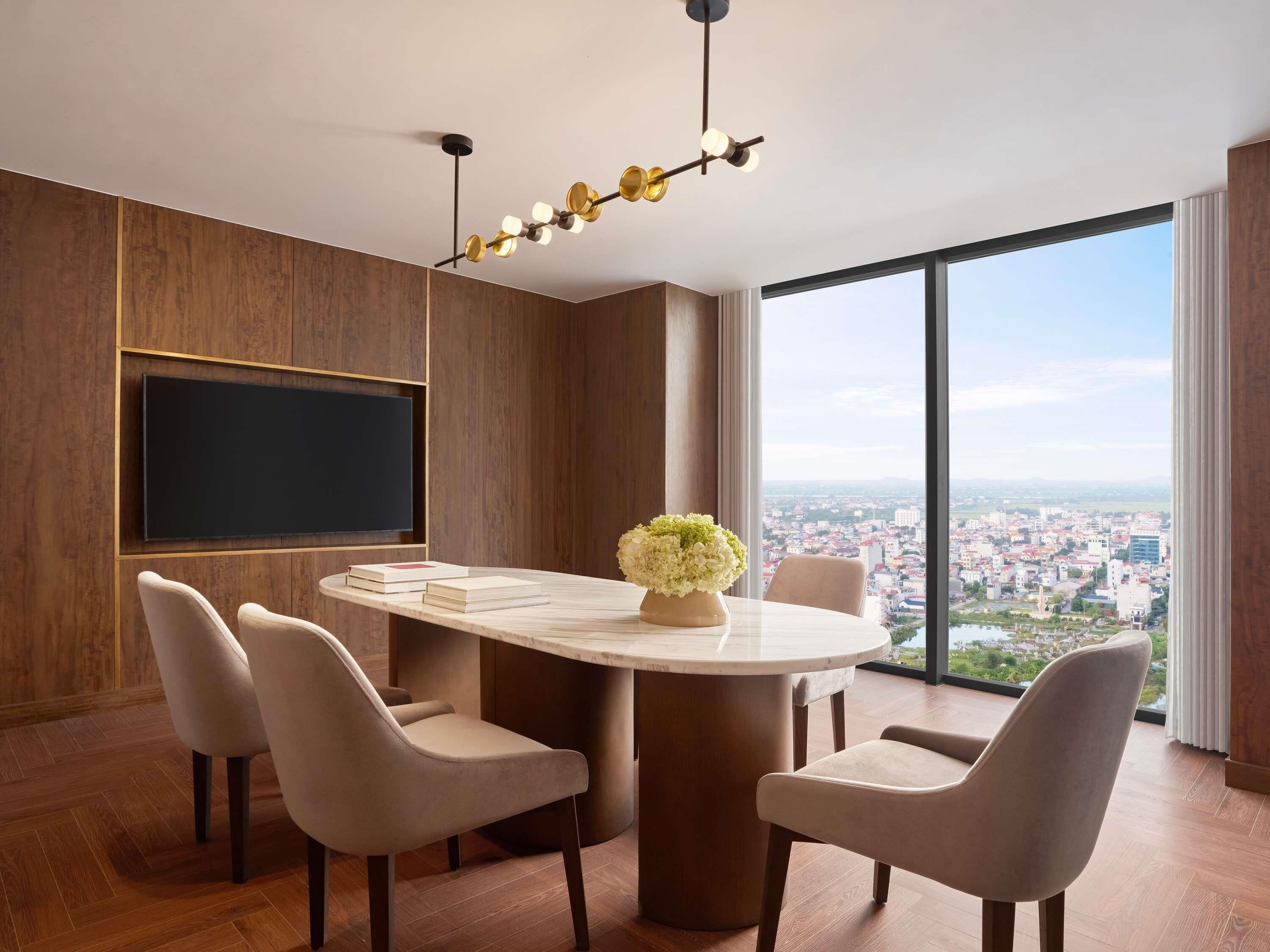 A modern dining room with a round table, four chairs, a large window with a city view, and a flat-screen TV mounted on the wall.