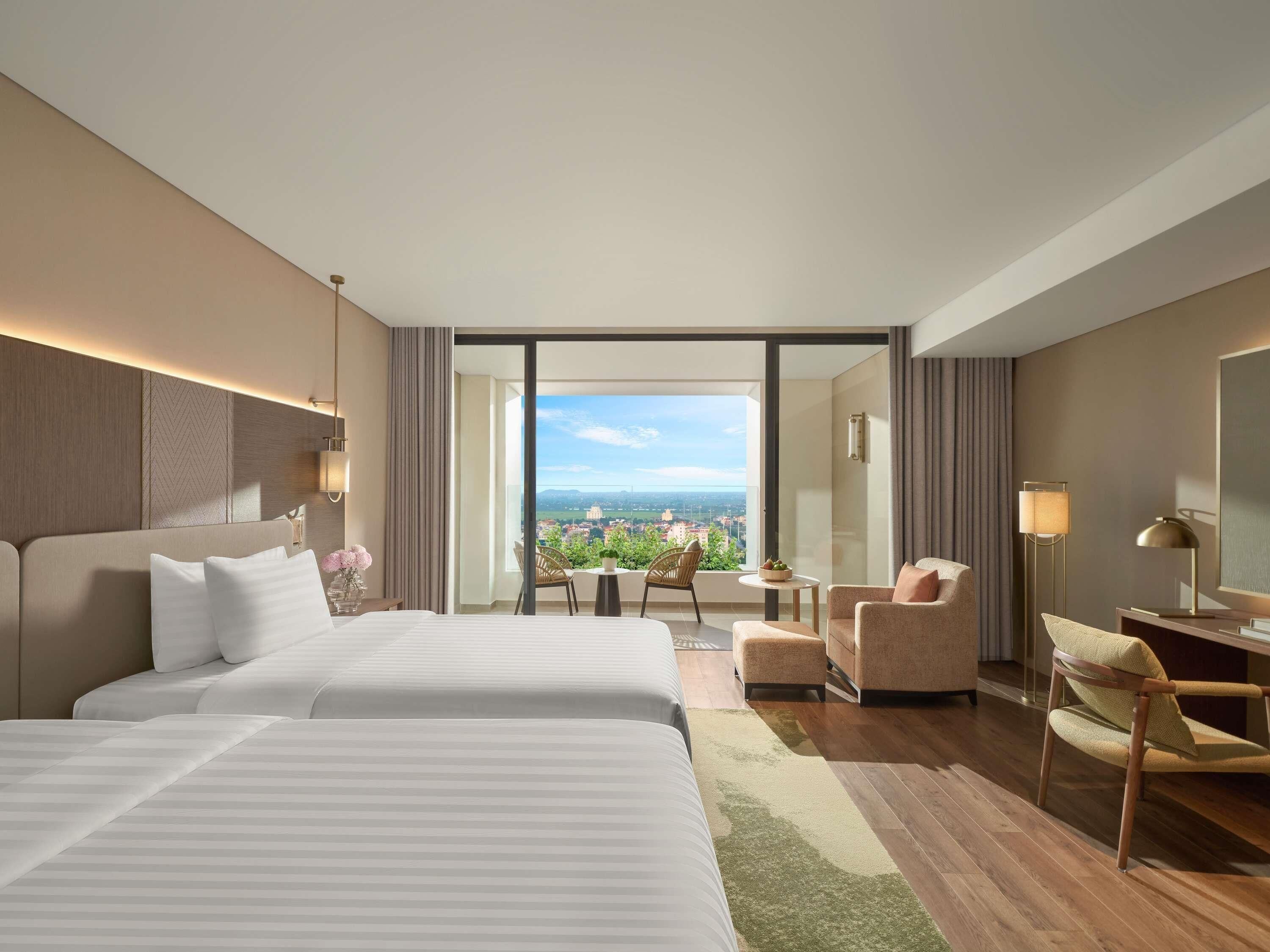 A hotel room with two beds, a balcony, a small table, a chair, and a lamp.