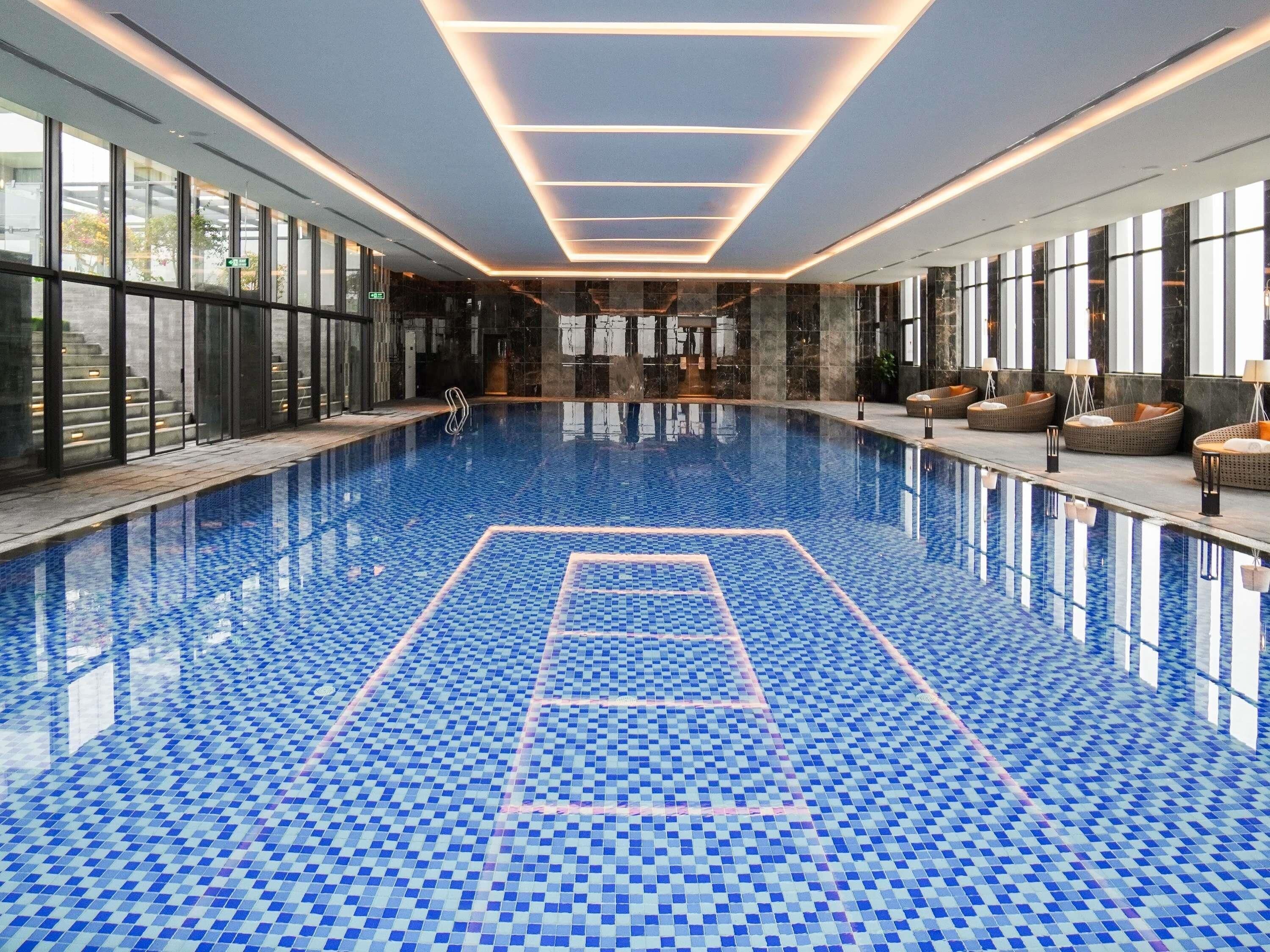 Ein großes Hallenbad mit Schachbrettmuster auf dem Boden, umgeben von Glaswänden und möblierten Sitzbereichen.