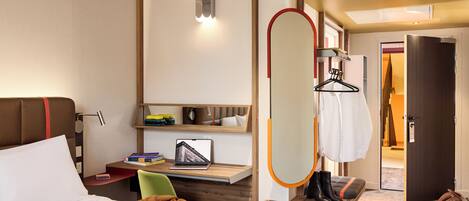 A hotel room with a bed, a desk with a laptop, a chair, a mirror, a coat rack, and a door leading to another room.