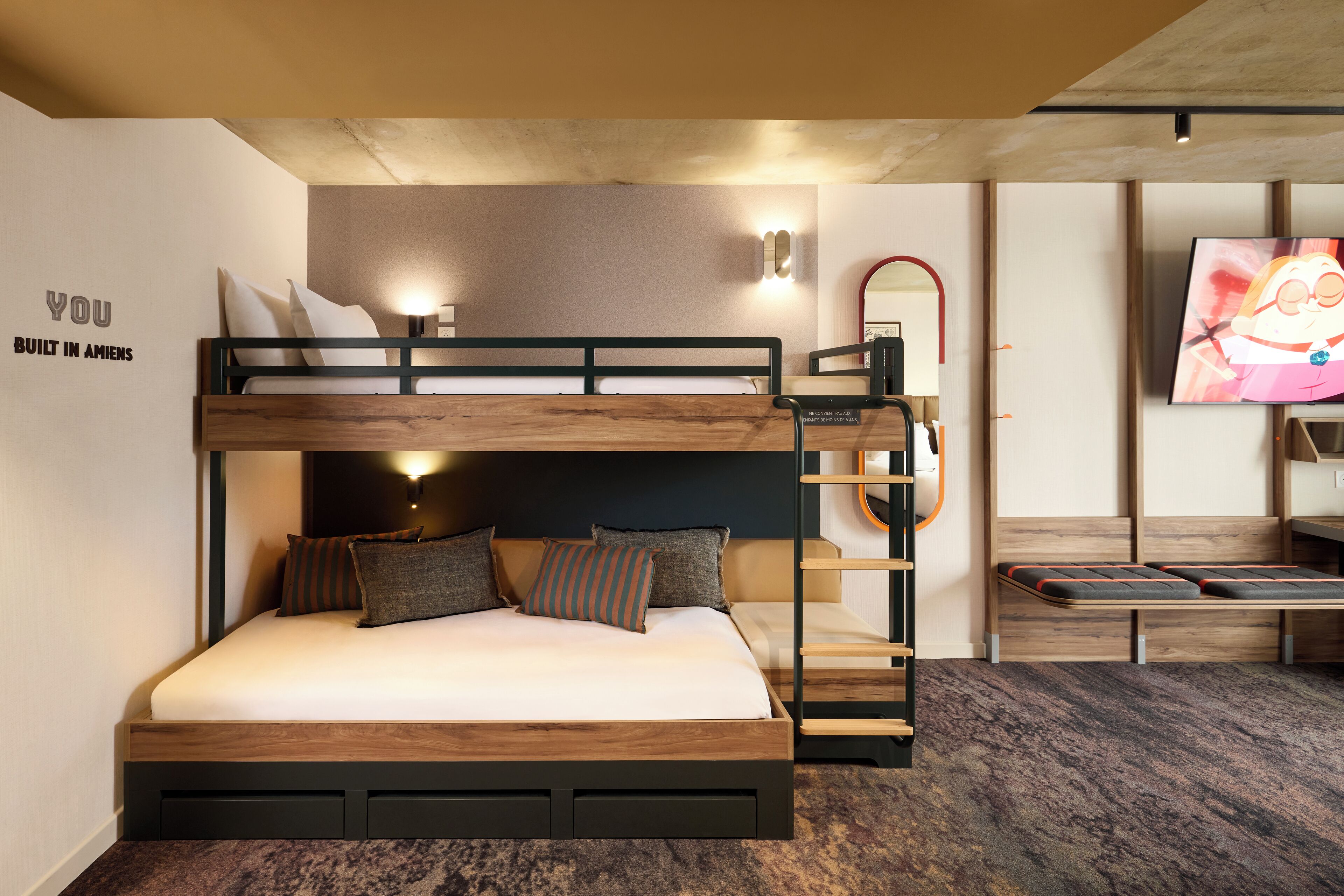 Una litera con estructura de madera, una cama inferior con almohadas, un escritorio pequeño, un espejo y un televisor en la pared.