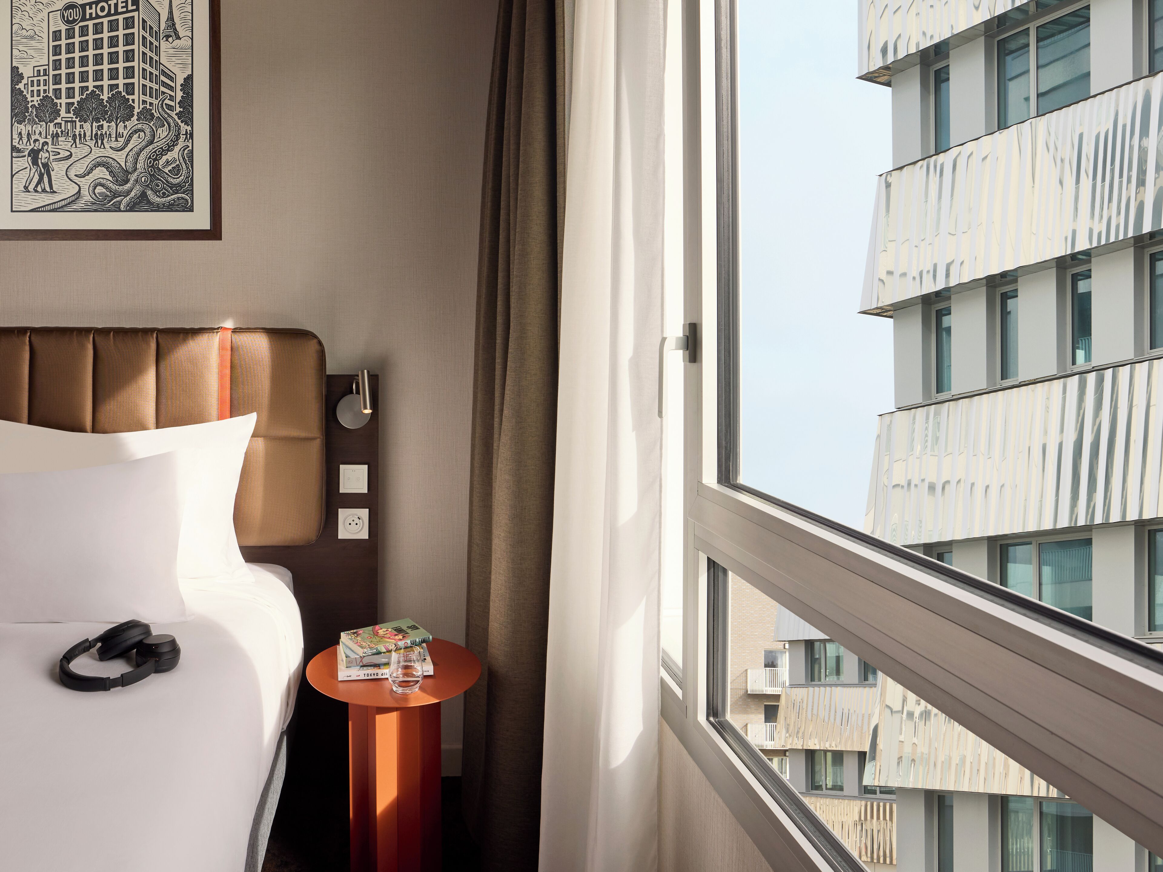 Habitación de hotel con cama, una mesita con un libro y un vaso, y una ventana con vista a un edificio.