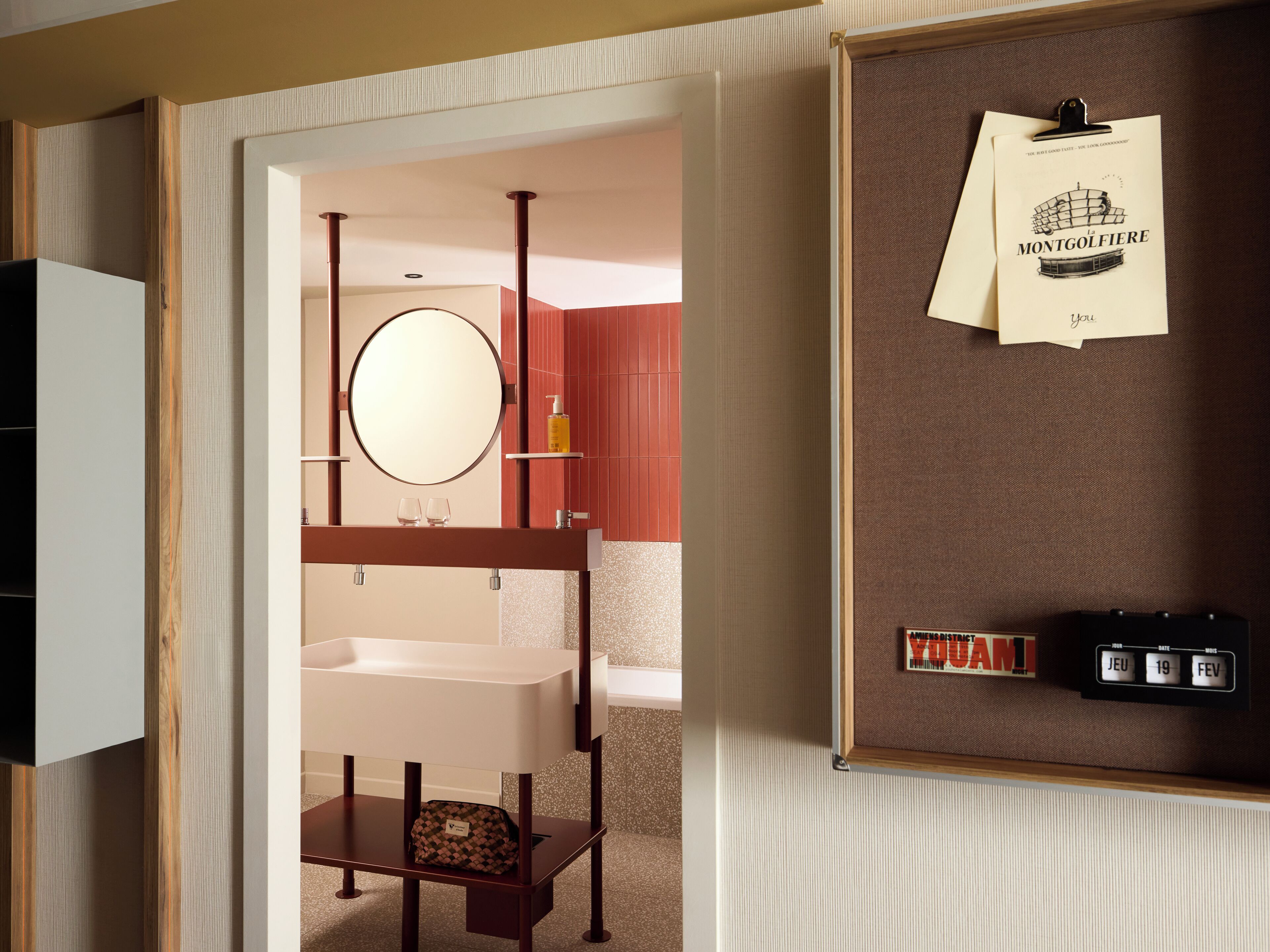 A modern bathroom with a wooden vanity, a round mirror, and a shelf with toiletries. A calendar and a framed picture are on the wall.