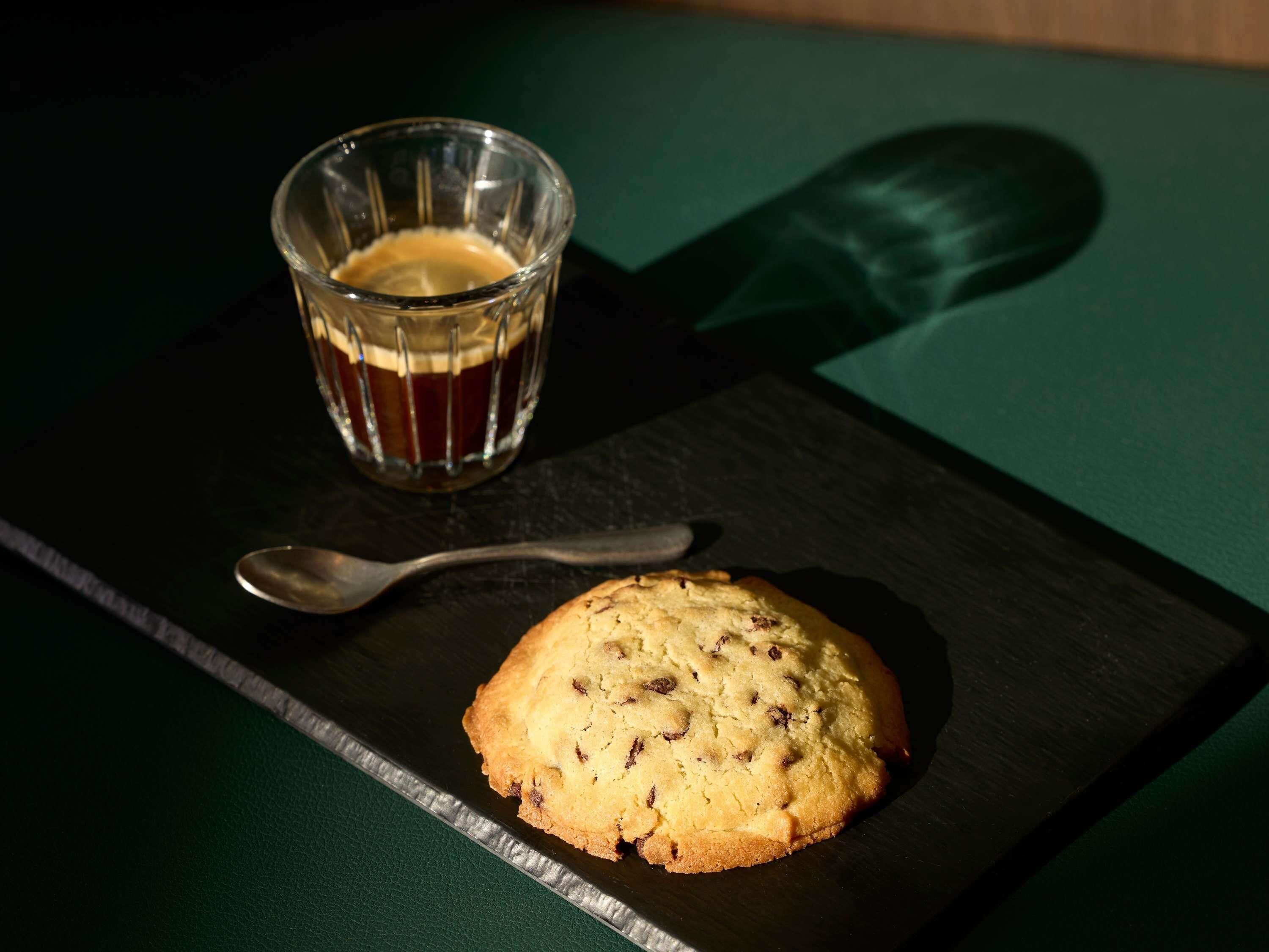 Una galleta sobre un plato de pizarra oscura con una cuchara a un lado, y un vaso de té sobre una mesa verde.