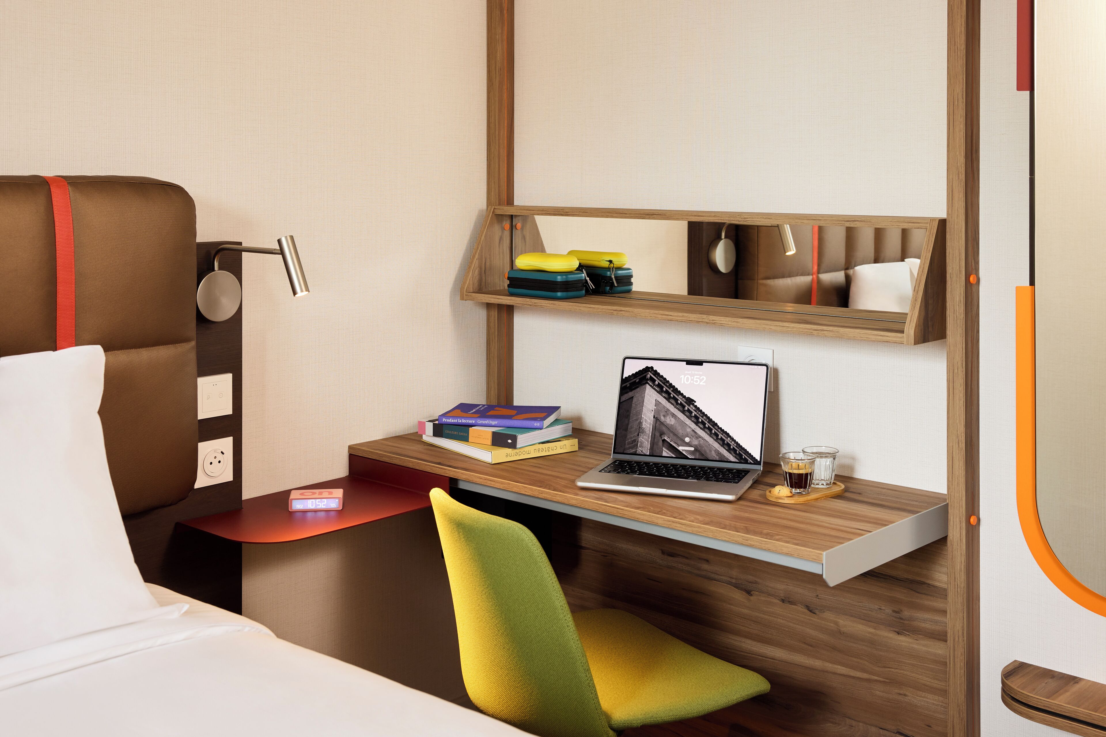 A modern hotel room with a bed, a desk with a laptop, books, and a chair.