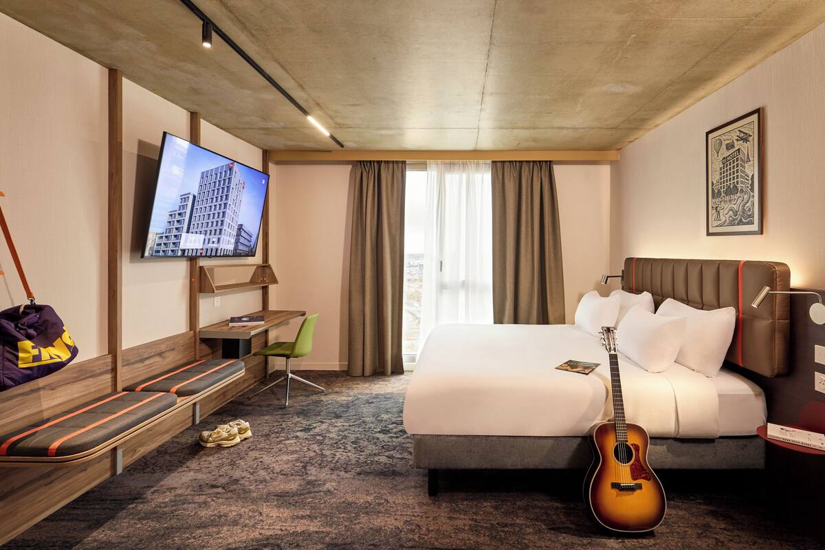Habitación de hotel con cama, televisor, escritorio, silla, una guitarra y vistas a la ciudad por la ventana.