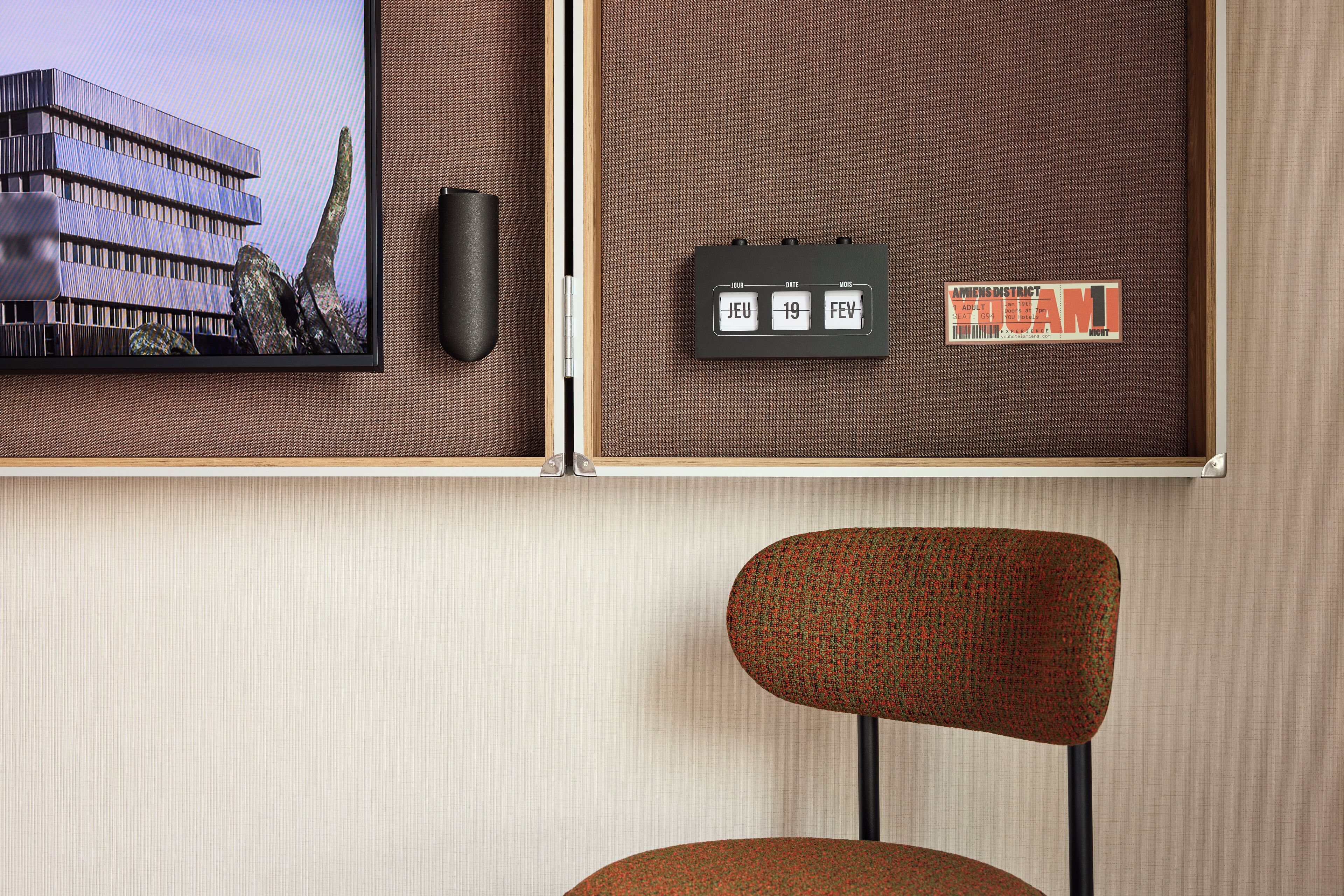A room with a mounted television displaying a building, a wall-mounted calendar, and a chair.