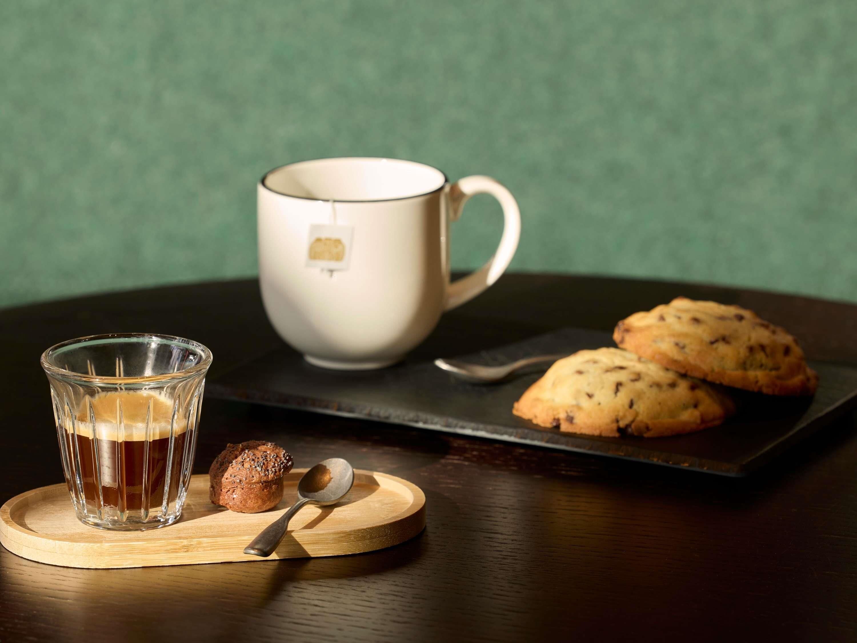 A glass of amber liquid, a white mug, and two cookies on a wooden coaster.