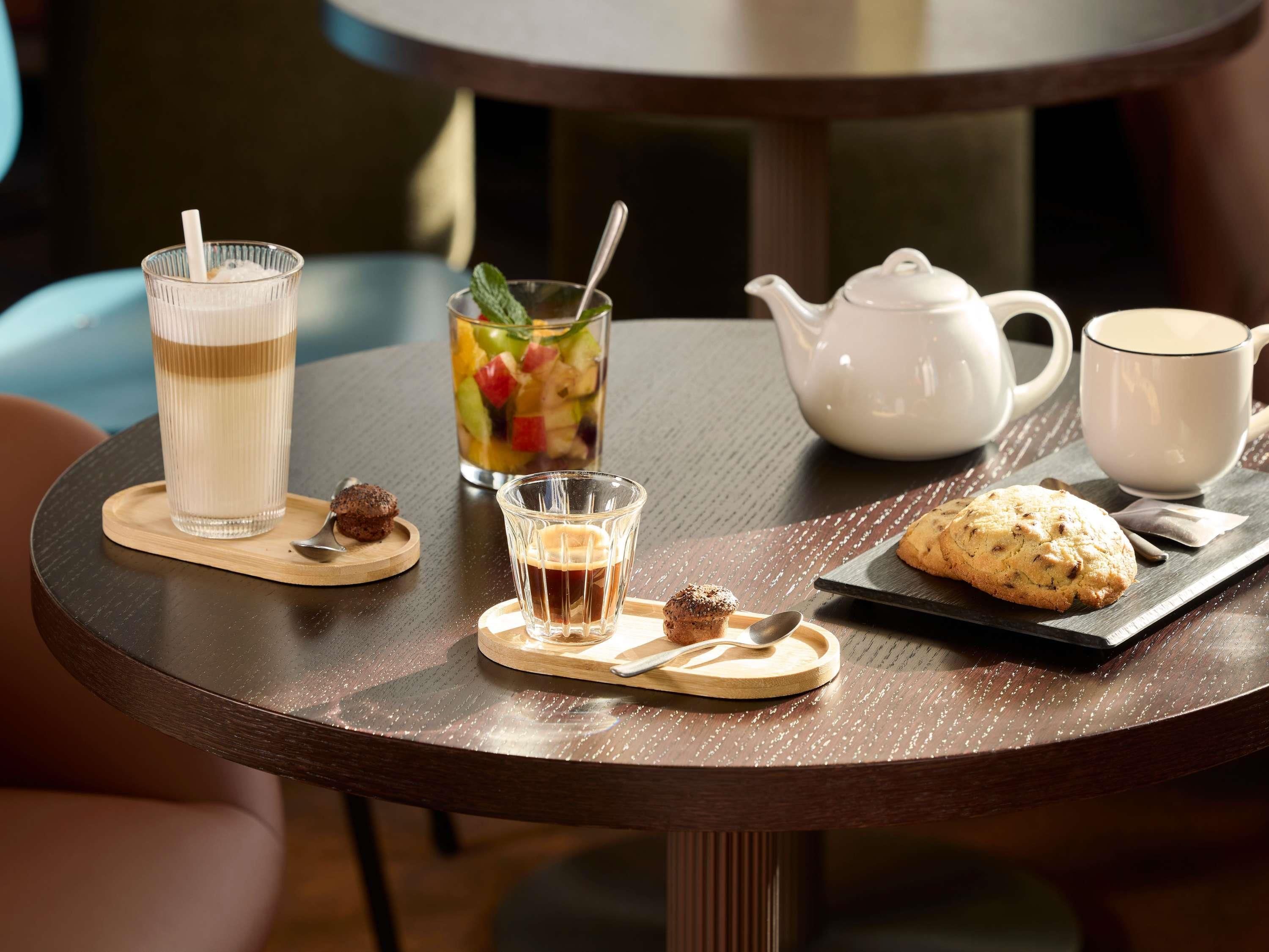 Een ronde tafel met een theepot, een kopje, een glas ijsthee, een glas koffie, een muffin en een fruitsalade.