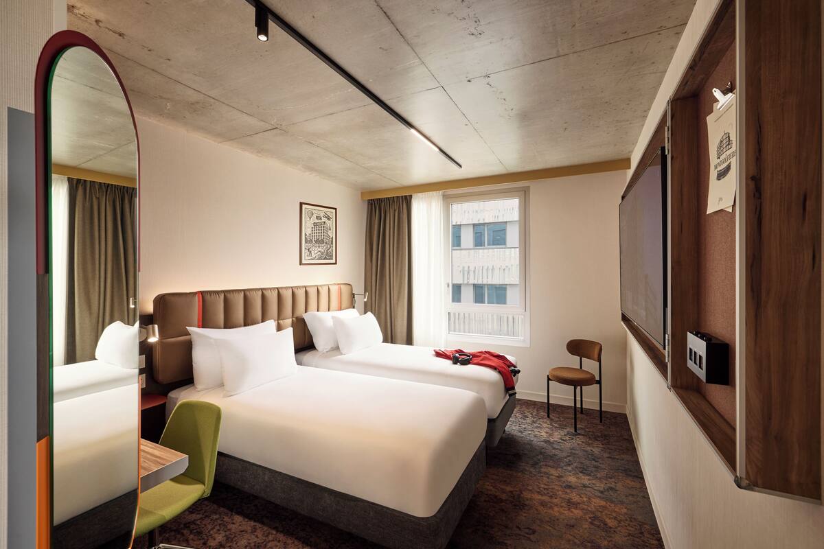 Habitación de hotel con dos camas, un escritorio, una silla, un espejo grande y una ventana con vistas a edificios.