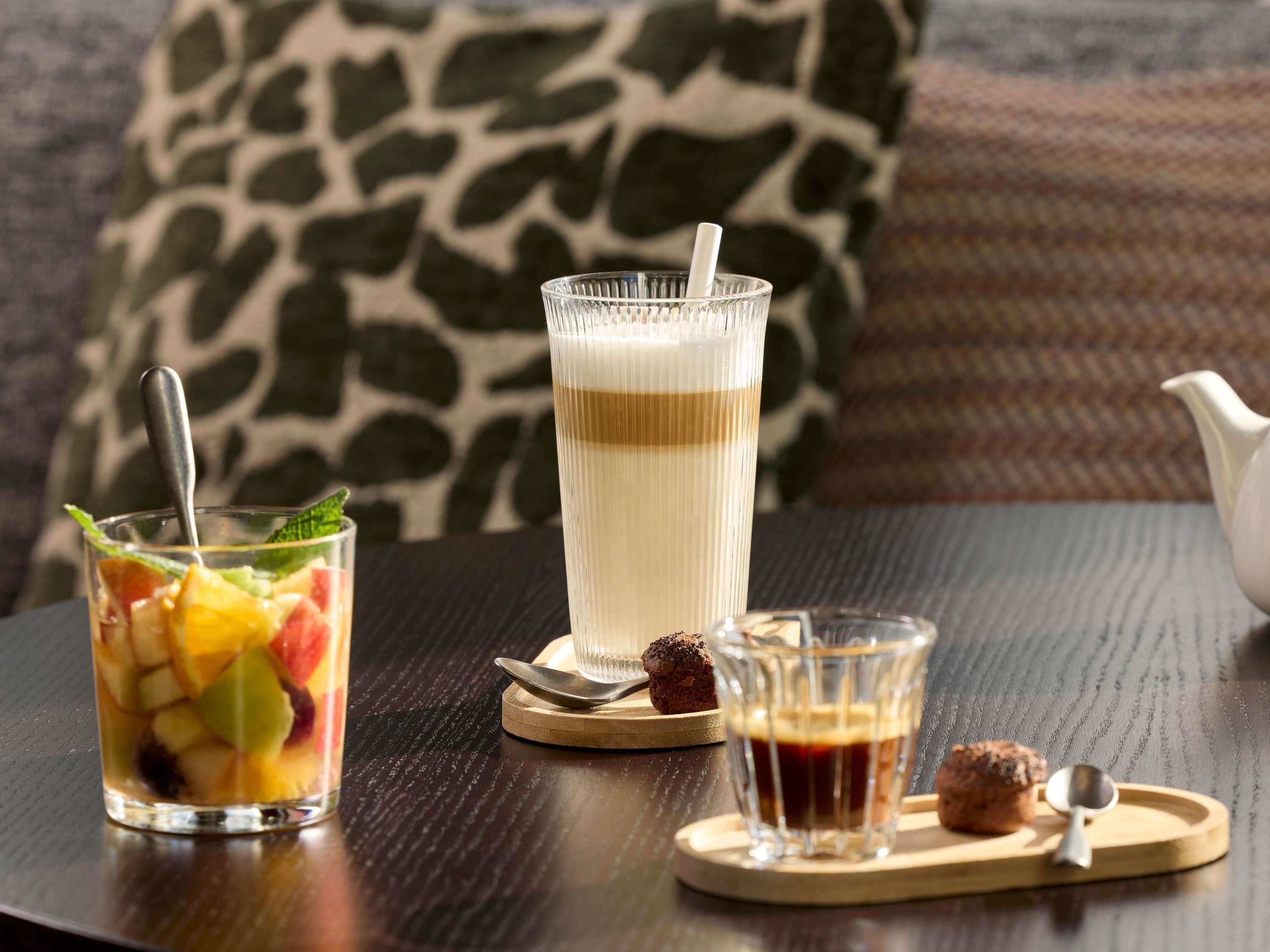 A table with a glass of iced coffee, a glass of fruit salad, and a muffin on a wooden coaster.