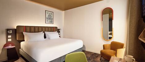 A hotel room with a large bed, a green chair, a small table with a cup, and a mirror on the wall.