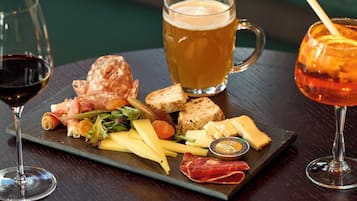 Una tabla de embutidos con queso, carnes y pan, acompañada de dos copas de vino y una jarra de cerveza.