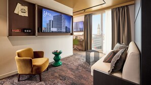 A hotel room with a TV mounted on the wall, a sofa, a chair, and a small table. - You Hotel Amiens - Handwritten Collection (Amiens)