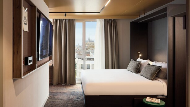A hotel room with a large bed, a TV mounted on the wall, a view of the cityscape through the window, and a small table with a book. - You Hotel Amiens - Handwritten Collection (Amiens)