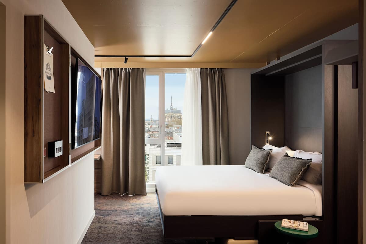 Una habitación de hotel con una cama grande, un televisor montado en la pared, vistas de la ciudad a través de la ventana y una mesita con un libro.