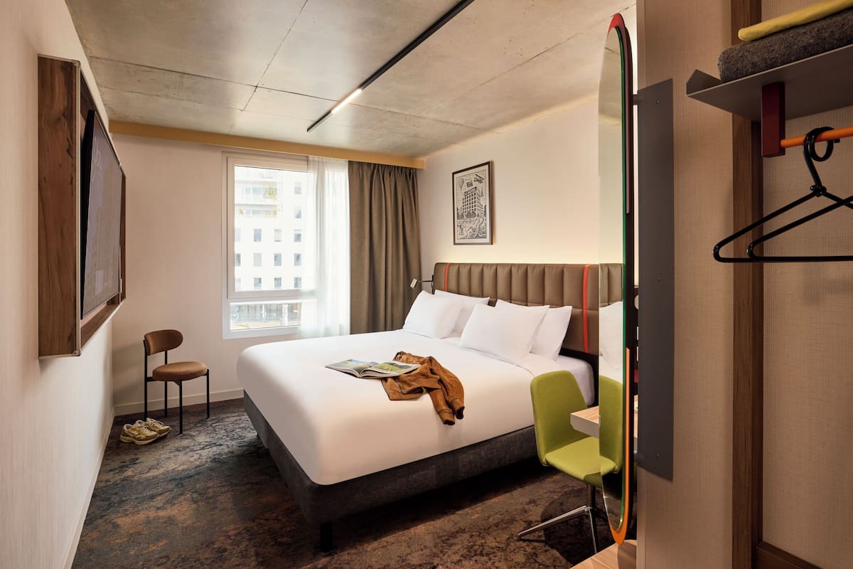 Habitación de hotel con una cama grande, una silla, un televisor y un ventanal con cortinas.