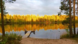 A sandy beach with a wooden dock extending into a calm lake, surrounded by trees reflecting golden autumn colors.