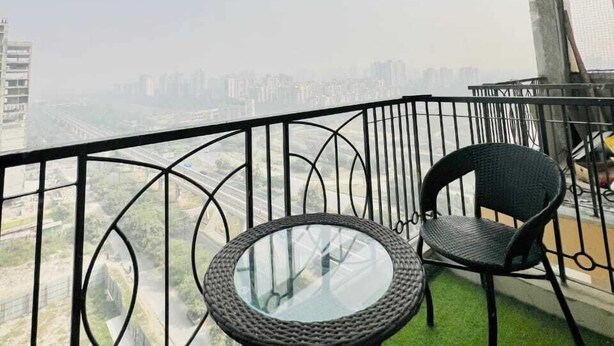 A balcony with a round glass table and two chairs, overlooking a cityscape. - High Sky In (Greater Noida)