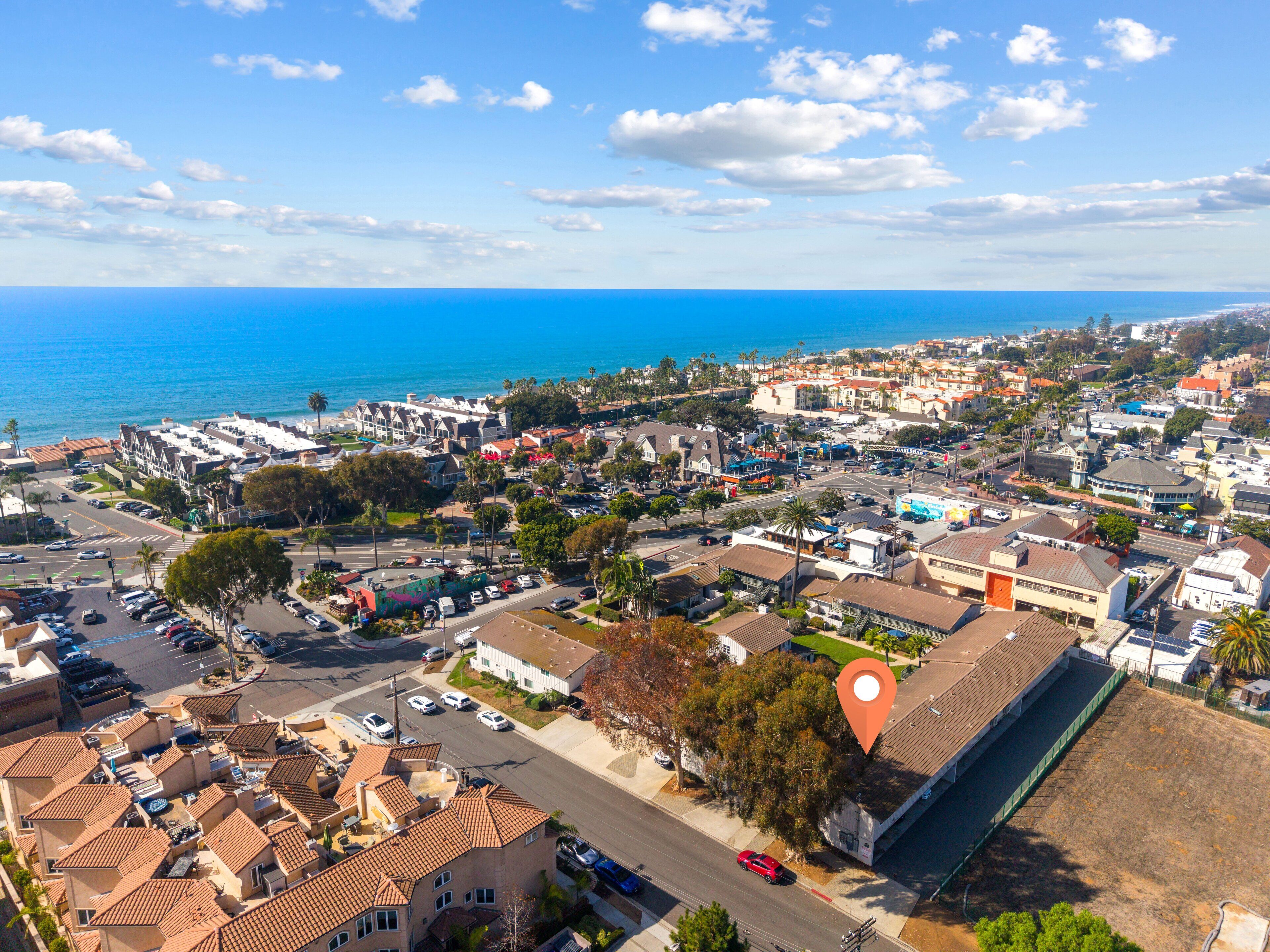 Une vue aérienne d’une ville côtière, avec un mélange de bâtiments résidentiels et commerciaux, un hôtel imposant doté d’un toit rouge, et une vue dégagée sur l’océan.