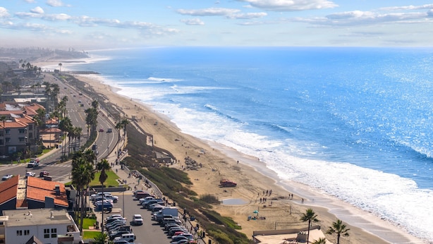 Una carretera costera con coches aparcados, una playa y vistas al océano.