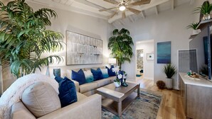 Una sala de estar moderna con un sofá color beige, cojines azules, una mesita de centro y plantas verdes grandes.