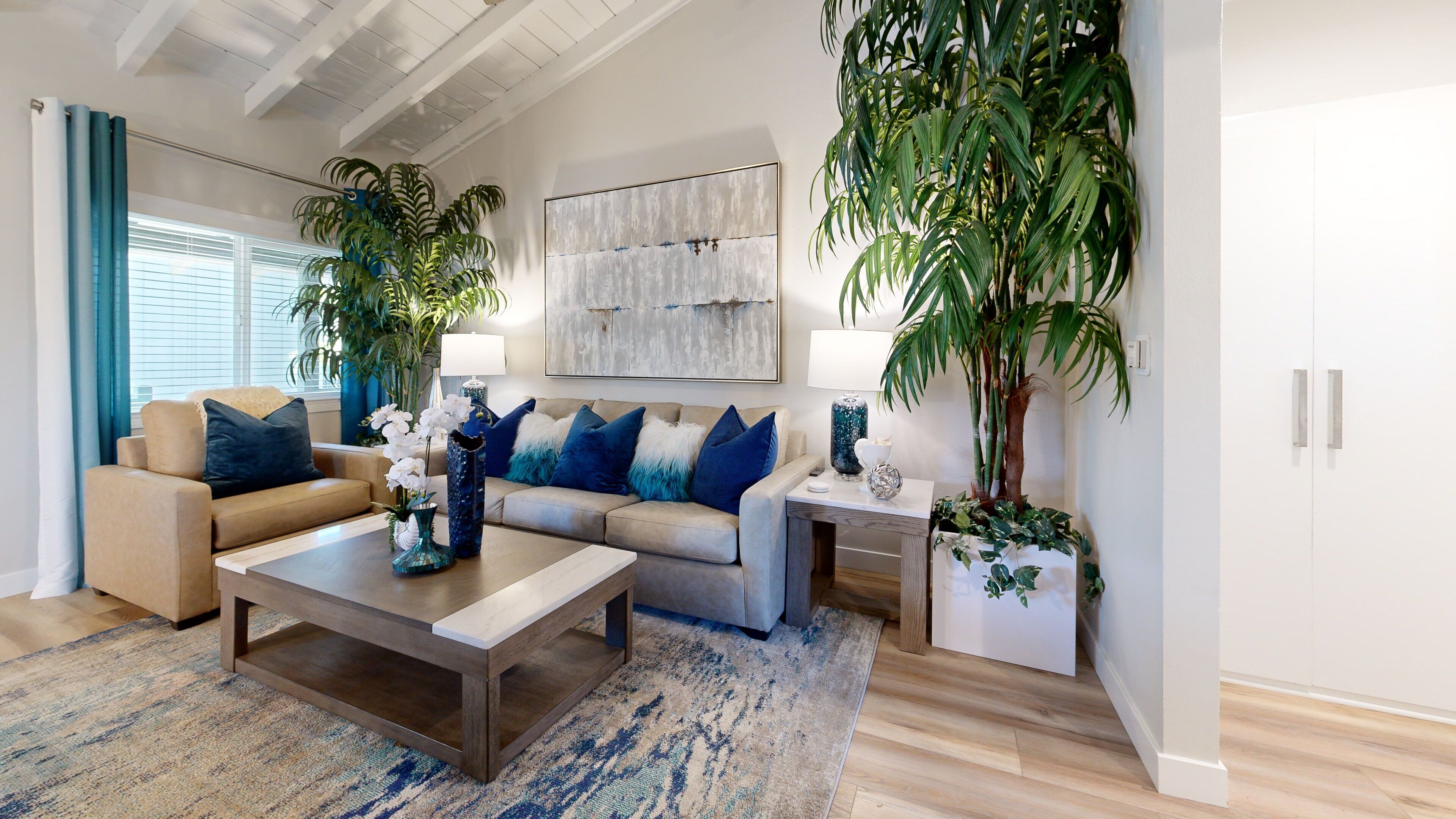 A modern living room with a sofa, coffee table, and two side tables. There are potted plants and a painting on the wall.