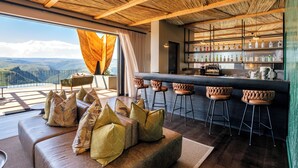 A modern bar area with a view of the mountains, a sofa with patterned cushions, and a bar counter with stools. - Mantis Hiddn In Addo (Gqebergha)