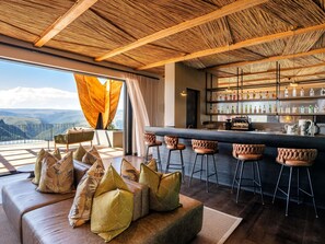 A bar area with a view of the mountains, a sofa with cushions, and a bar counter with bottles. - Mantis Hiddn In Addo (Gqebergha)