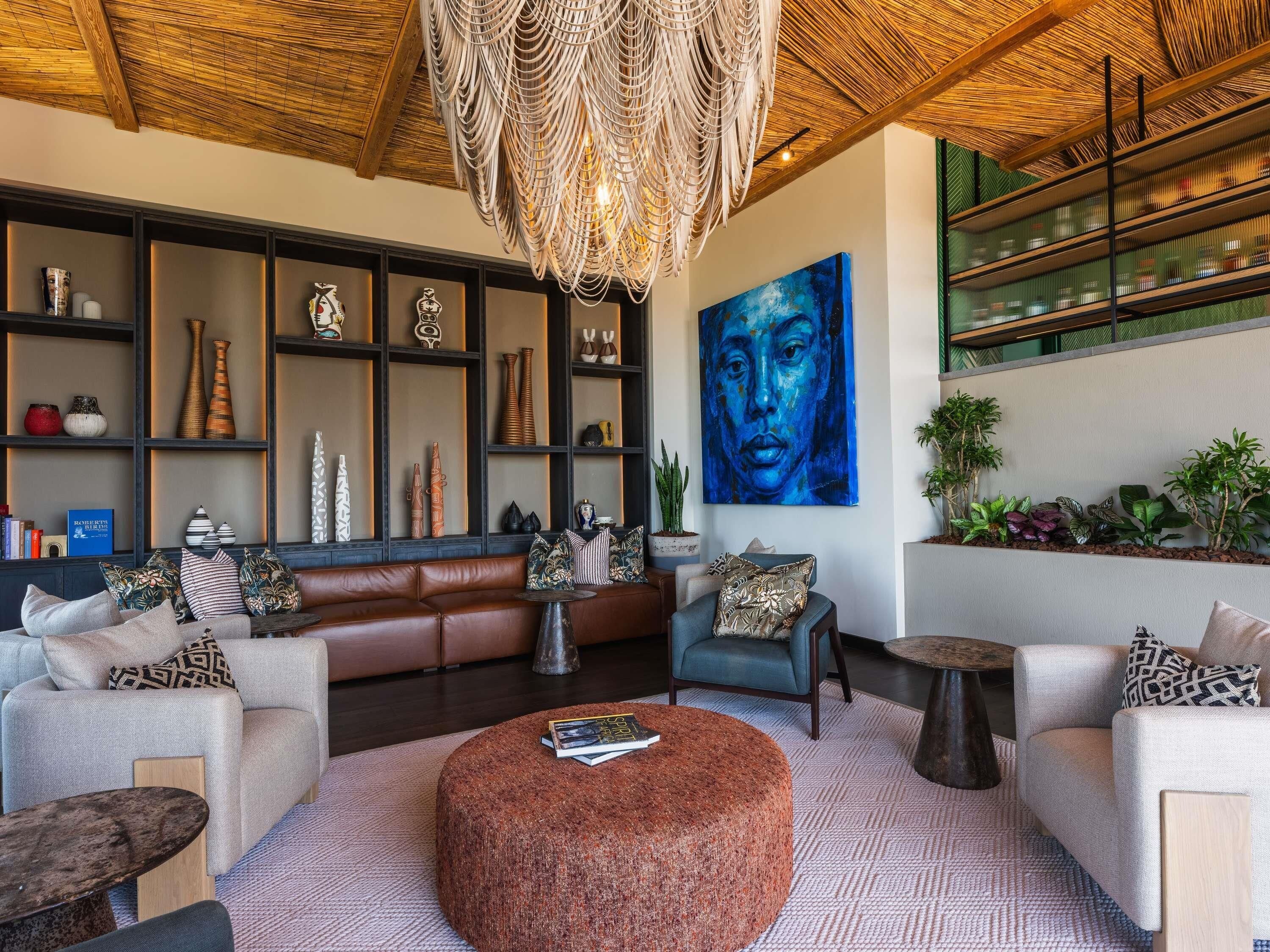 A modern living room with a brown leather sofa, a round ottoman, and a blue armchair. There is a large abstract painting on the wall and a variety of decorative items on shelves.