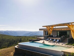 A modern outdoor pool area with a view of distant mountains, a wooden pergola, and a seating area with lounge chairs. - Mantis Hiddn In Addo (Gqebergha)