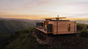 A wooden cabin with a balcony overlooking a mountainous landscape during sunset. - Mantis Hiddn In Addo (Gqebergha)