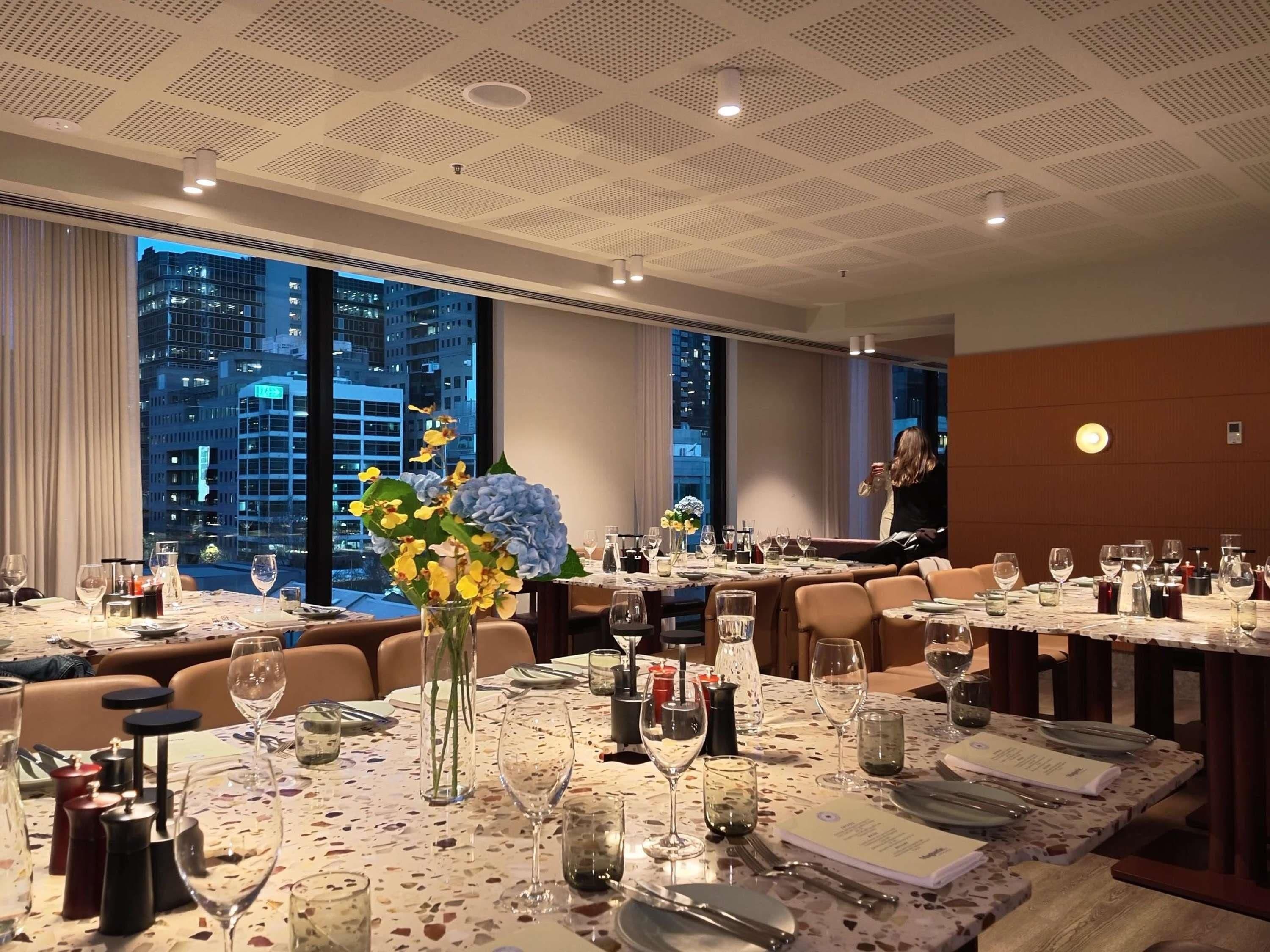 Un restaurante con varias mesas dispuestas para comer, amplias ventanas que ofrecen vistas a la ciudad y un techo con diseño de cuadrícula.