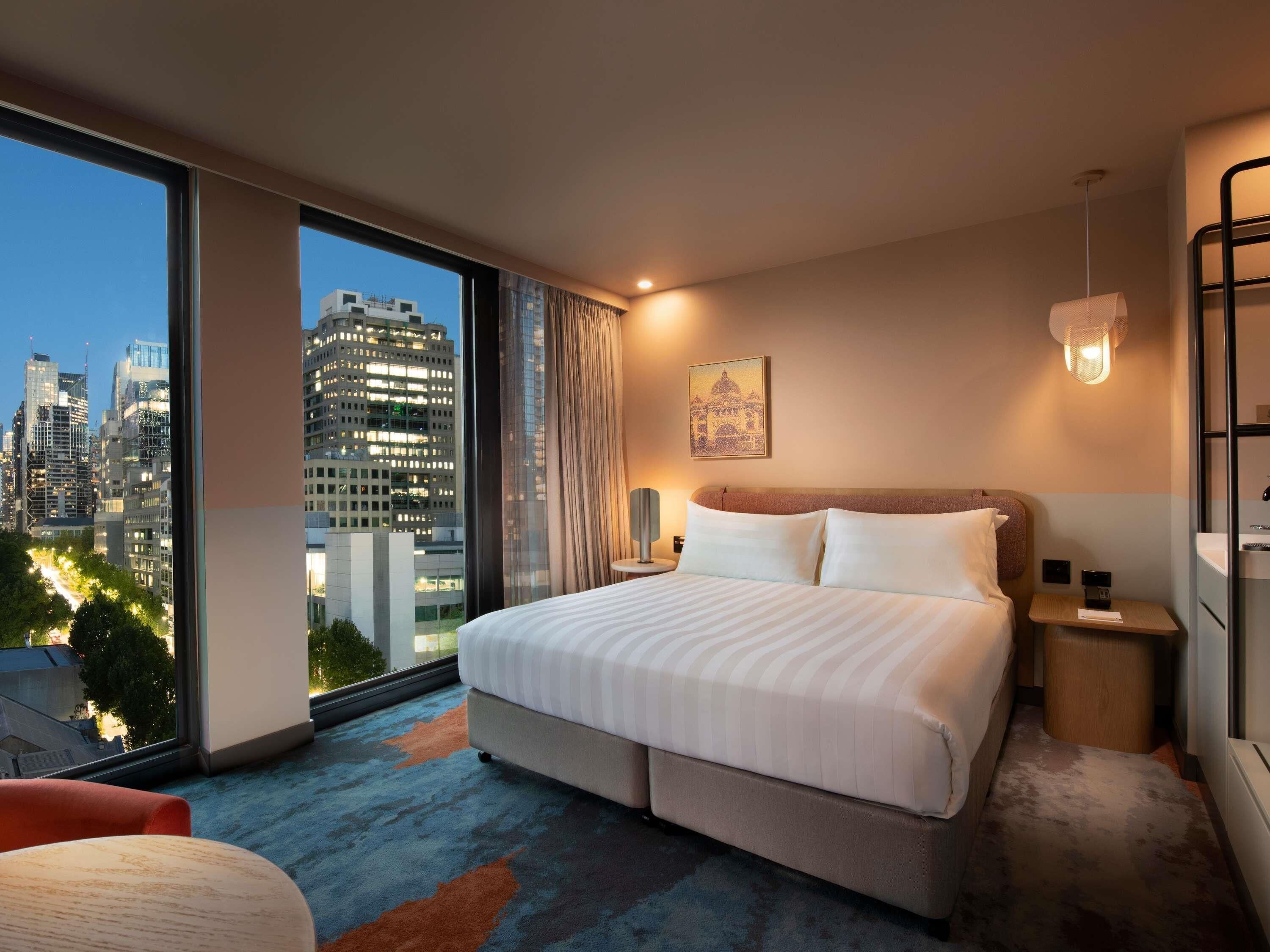 Une chambre d’hôtel avec un grand lit, une table de chevet, une lampe et une vue sur le paysage urbain.