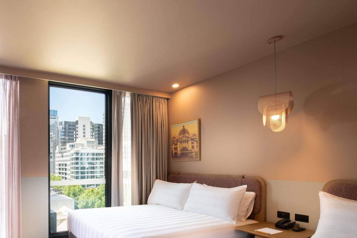 A hotel room with a large window, a bed, a nightstand, a lamp, and a painting on the wall.