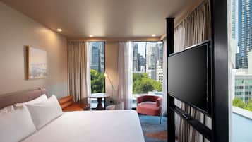 Quarto de hotel com uma cama grande, uma mesinha, uma cadeira e uma televisão.