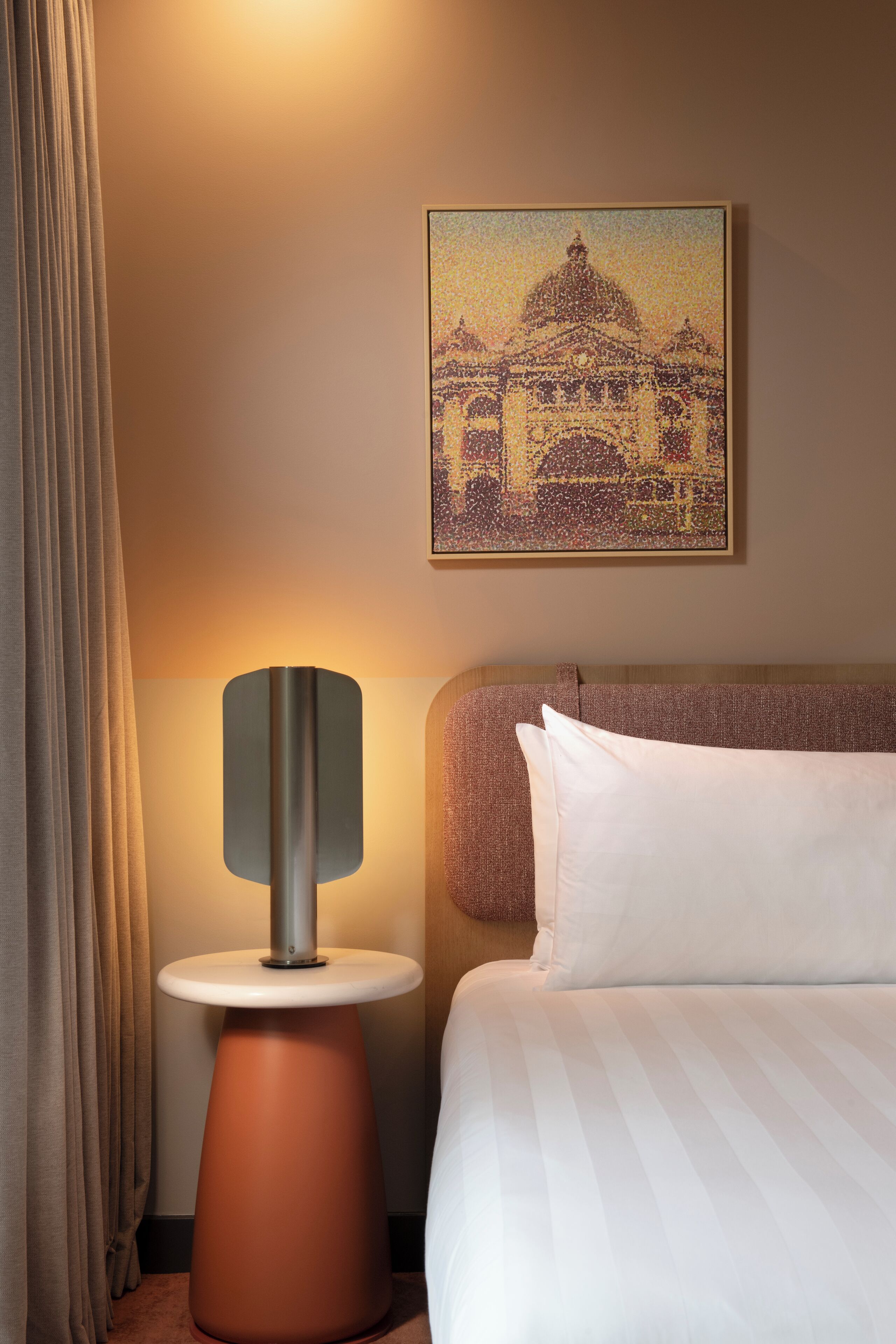 Ein Hotelzimmer mit einem Bett, einem Nachttisch mit Lampe, einem Bild eines Gebäudes und einem Vorhang.