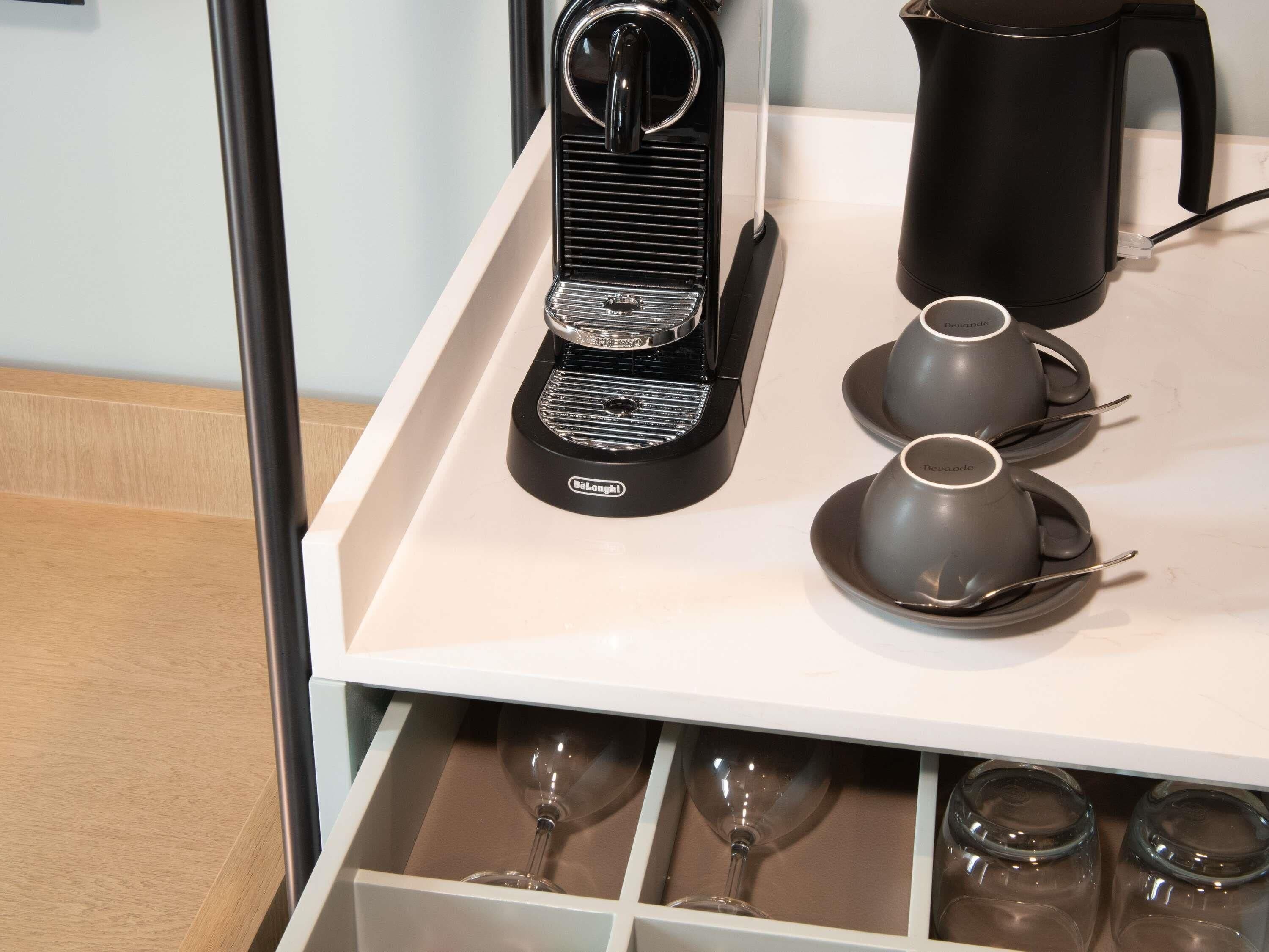 A kitchen counter with a coffee maker, two cups, a kettle, and a wine glass drawer.