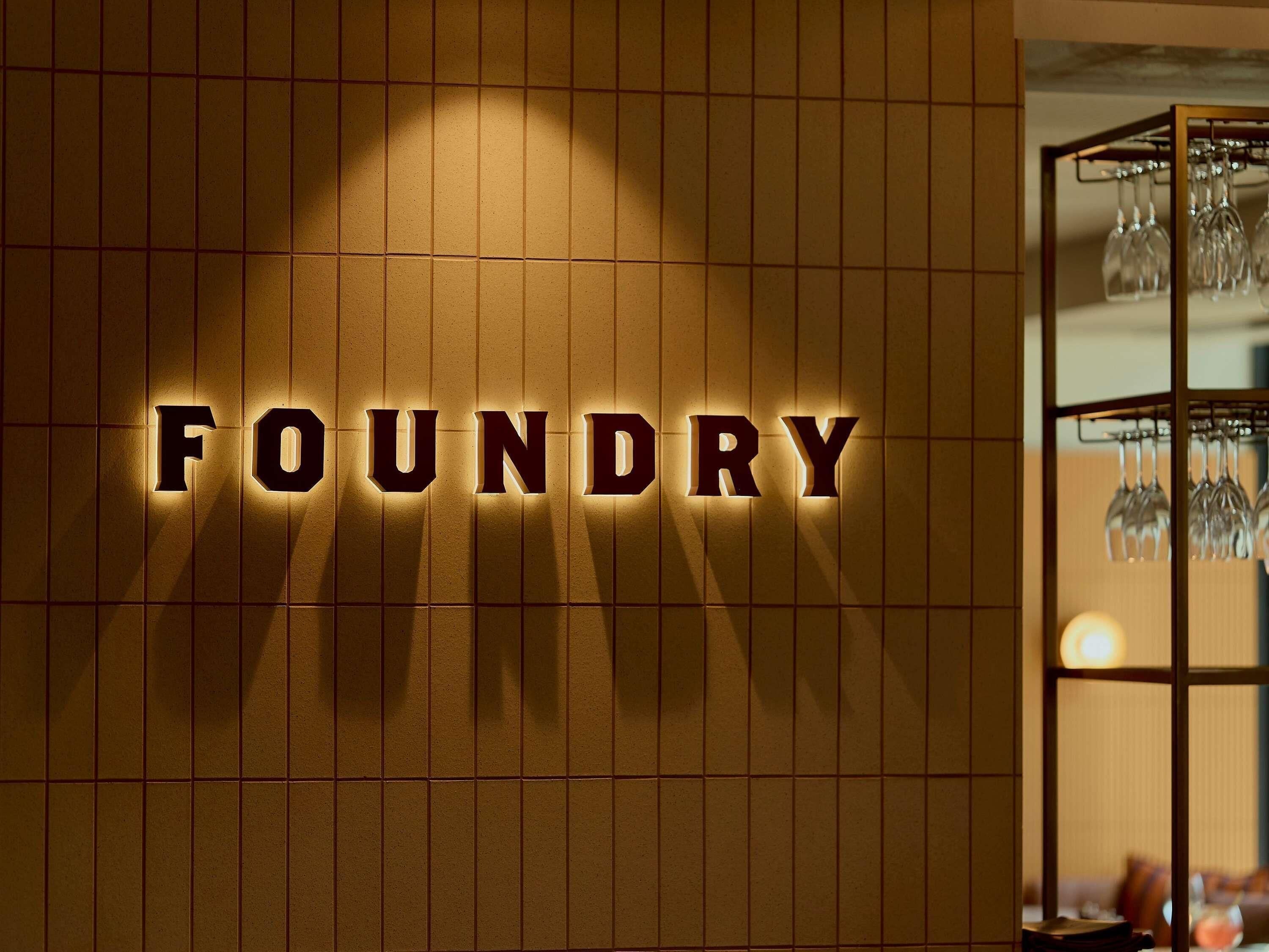 Una pared con letras iluminadas que dicen 'FOUNDRY' y un porta copas de vino al fondo.