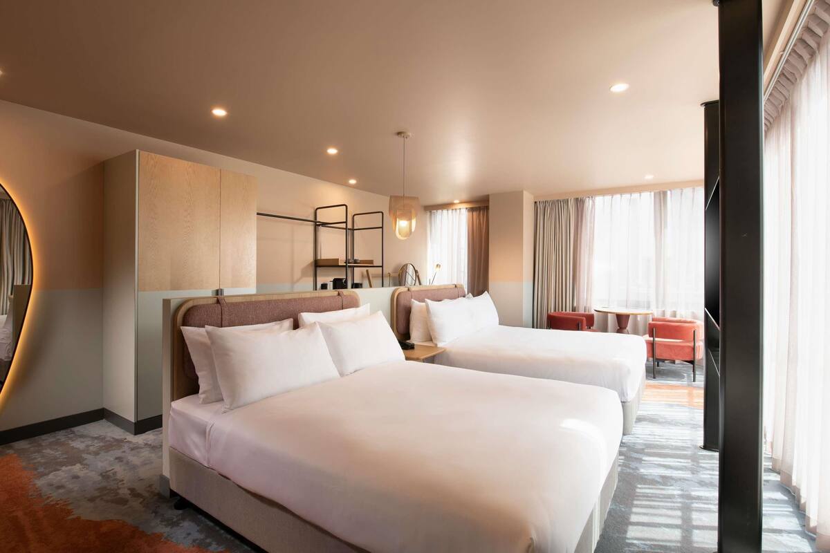 A hotel room with two beds, a sitting area with a table and chairs, a large mirror, and a view of the outdoors.