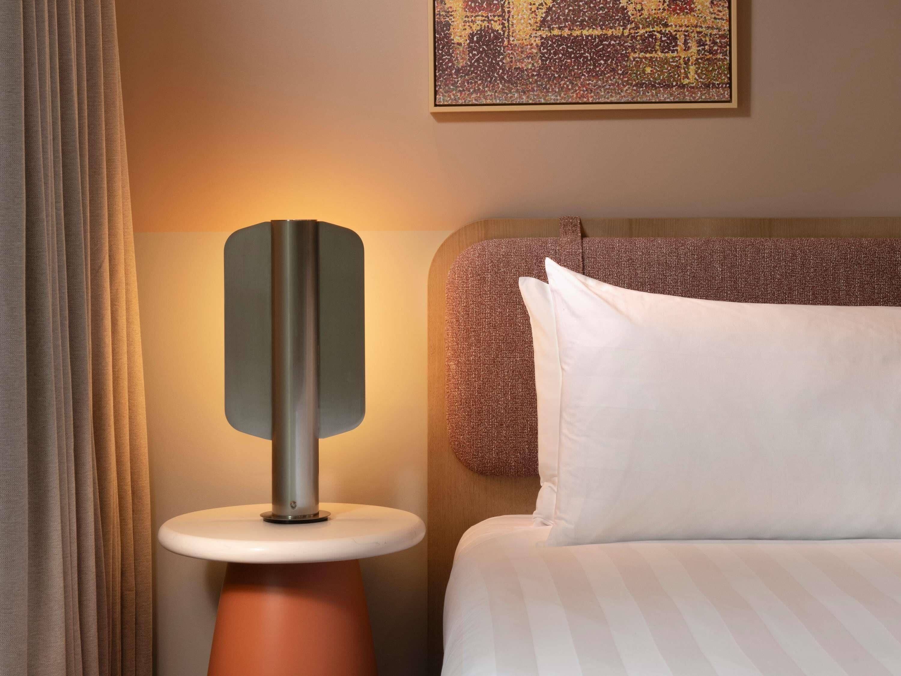 Une chambre d’hôtel avec un lit, une table de chevet avec une lampe, un tableau au mur et une fenêtre avec des rideaux.