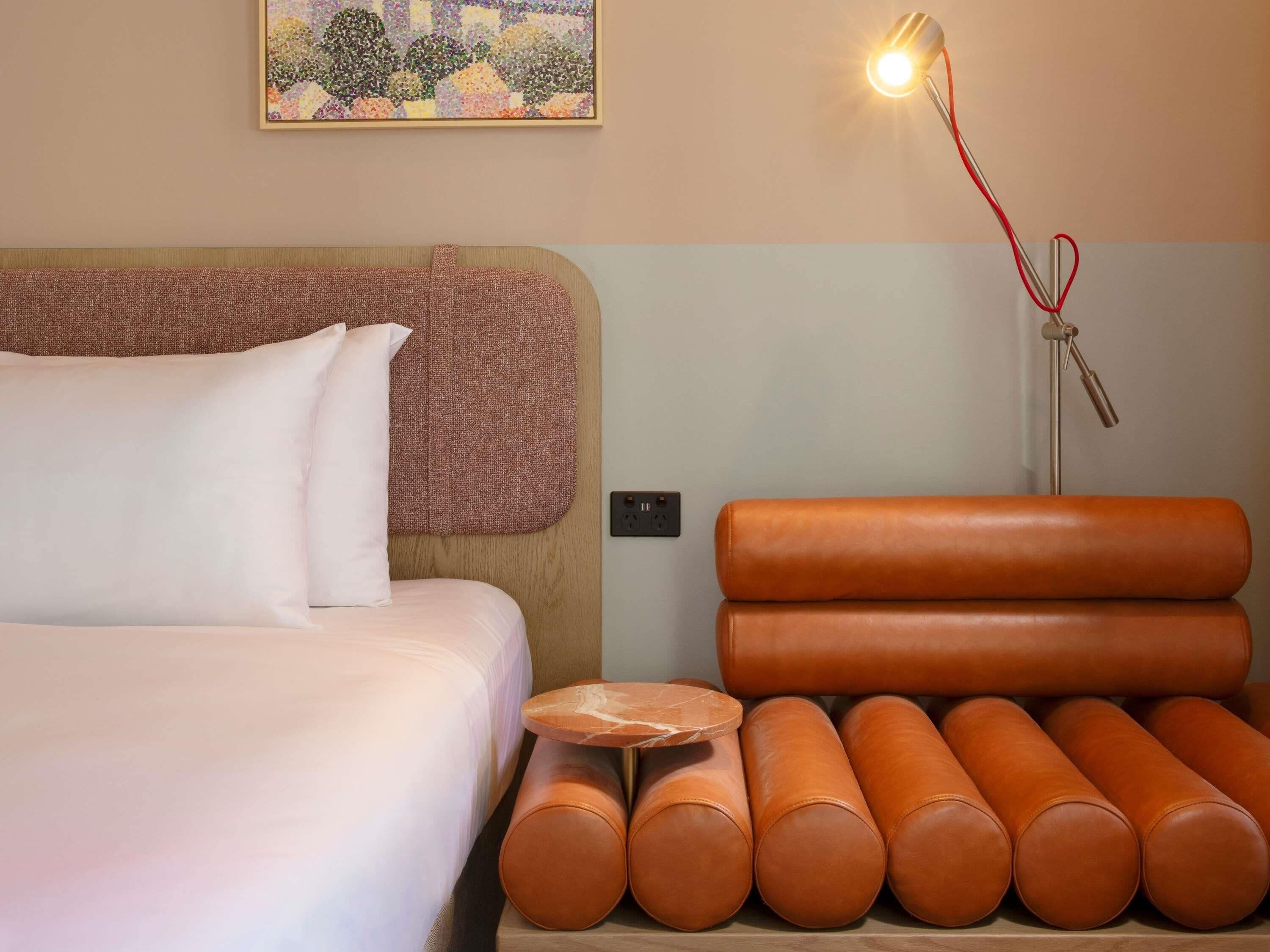 A neatly arranged hotel room with a bed, pillows, a headboard, a nightstand with a lamp, and a stack of cylindrical cushions.