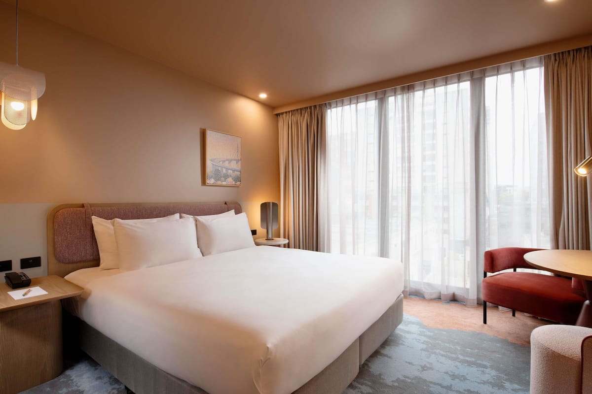 A hotel room with a large bed, a desk, a chair, and a view of the city through the window.